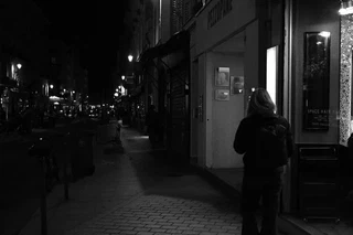 Black-and-white night street scene with a figure walking beside lit storefronts
