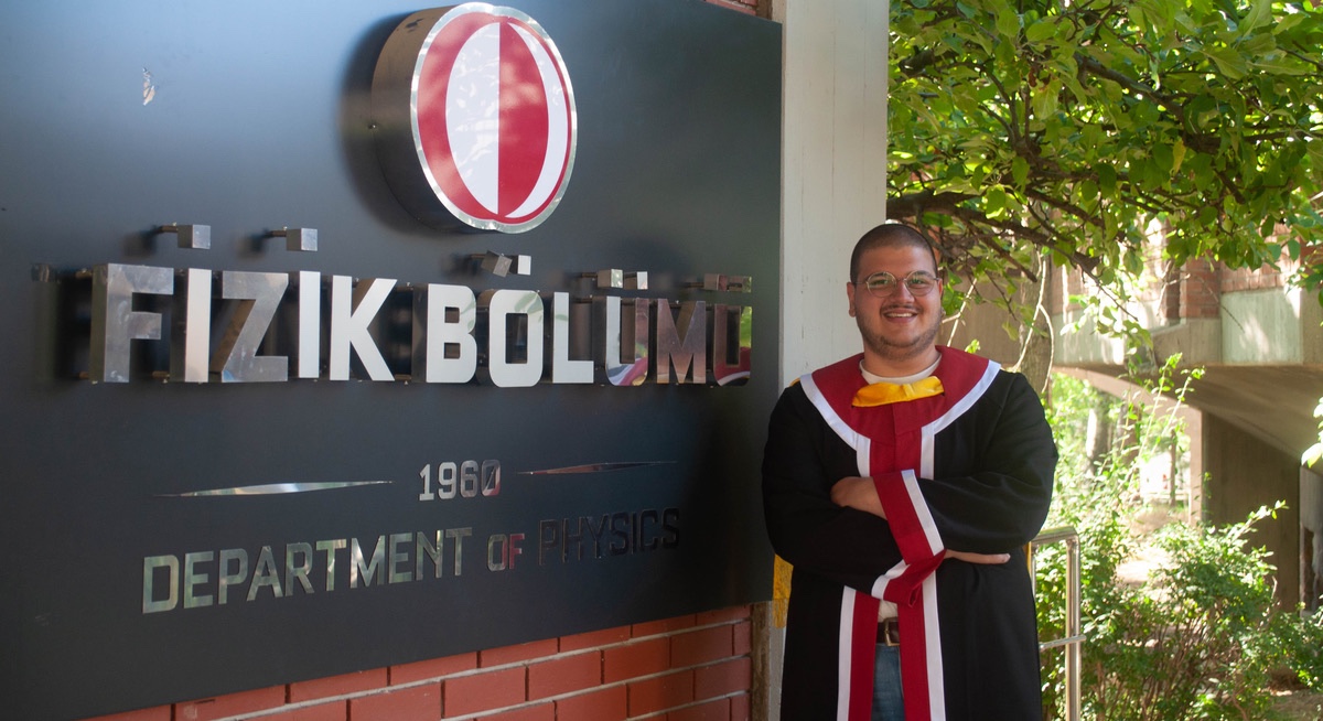 Ege standing beside the Department of Physics sign at graduation